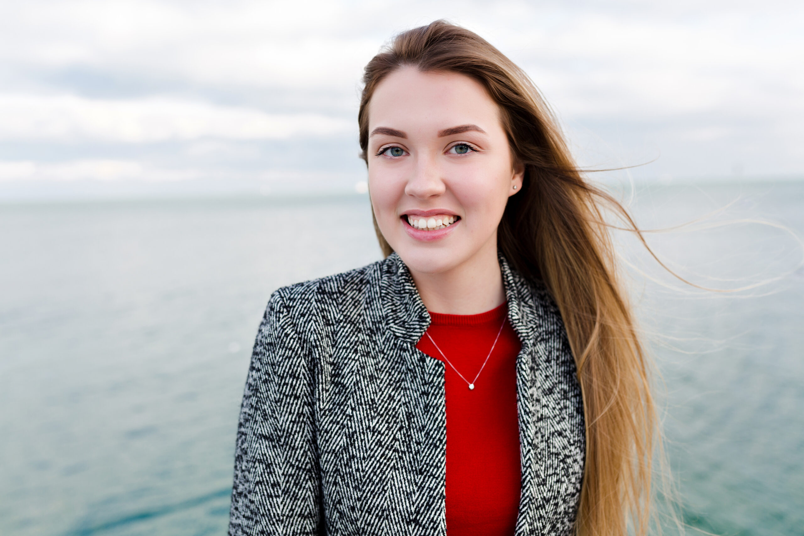 Happy woman smiling with long hair, blue eyes, wearing a red shirt and grey coat near the sea